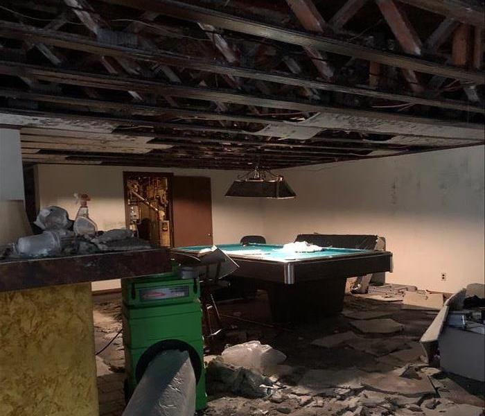 Charred ceiling showing rafters, debris on the floor by the pool table, and the green air scrubber with a duct at work